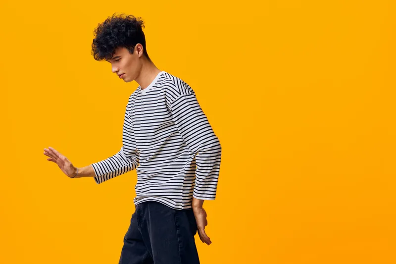 young man in striped shirt walking casually against bright orange background with curly hair, relaxed posture, and modern casual style