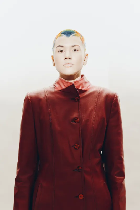 Woman with colorful short hair wearing a red coat stands confidently against a white background, showcasing modern fashion and bold style in a minimalistic setting.