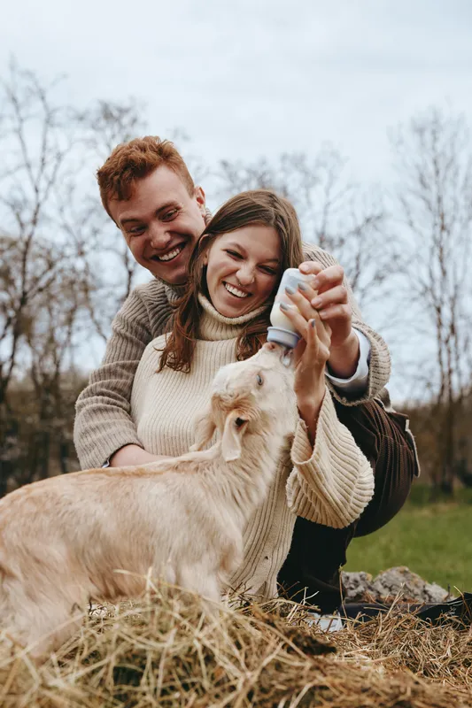 Young couple feeds a baby goat with a bottle outdoors in autumn while smiling and enjoying a cozy lifestyle moment together in warm sweaters.