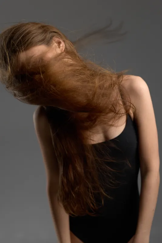 Woman with long brown hair flipping hair wearing black sleeveless top, motion blur effect in studio lighting.