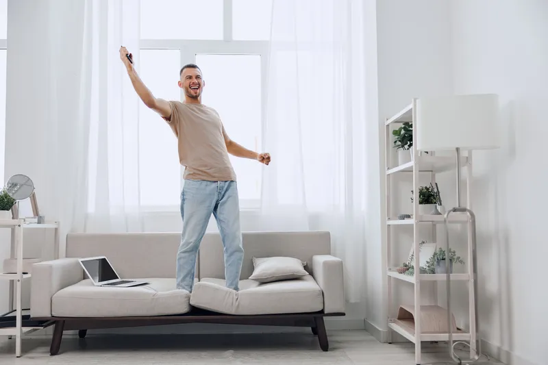 Joyful man celebrating at home, standing on a couch with a remote in hand, vibrant decor and bright natural light enhancing the lively atmosphere.