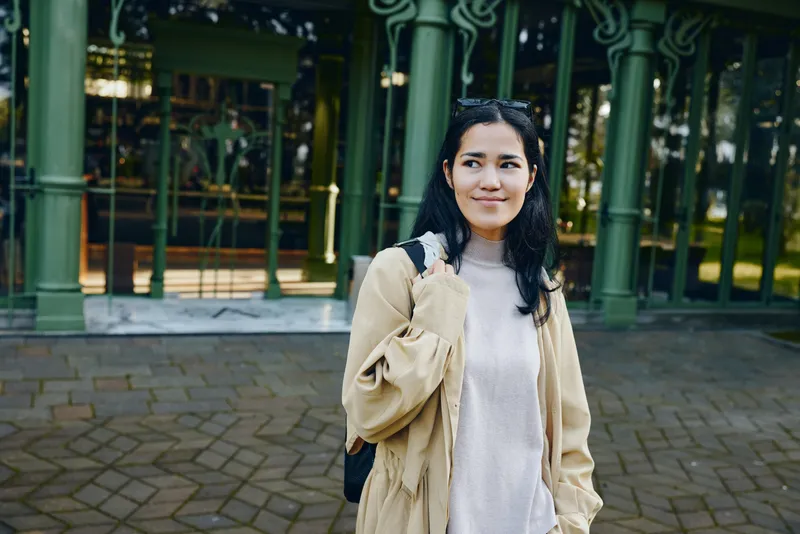 Woman in a casual beige coat and light sweater stands outdoors near a glass building, relaxed smile, everyday street style, natural light, candid moment, urban lifestyle.