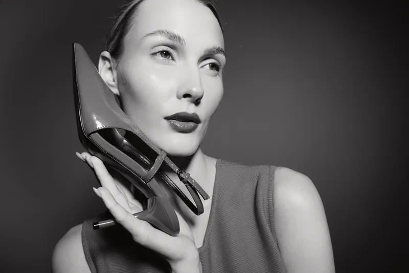 Caucasian woman with light skin holds a high heel shoe near her face, looking away. Black and white fashion portrait with elegant and stylish concept.