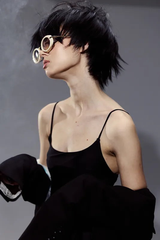 Young woman in stylish glasses and black outfit posing with moody expression against gray background