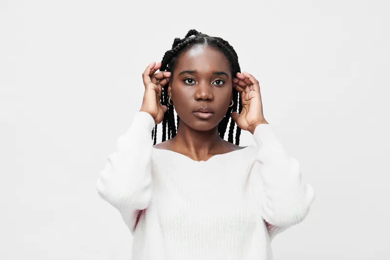 A young African woman with braided hair, expressing calm confidence while adjusting her hair in a cozy white sweater against a minimalist light background