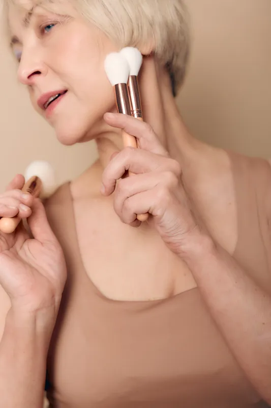 Mature woman with short blonde hair applies makeup brushes on face and neck, wearing beige top. Concept of beauty routine, skincare, self-care, and natural aging with soft neutral background.
