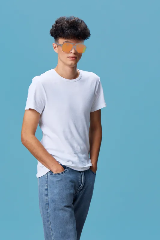 young man wearing orange sunglasses standing casually with hands in pockets against plain blue background in stylish white t-shirt and jeans