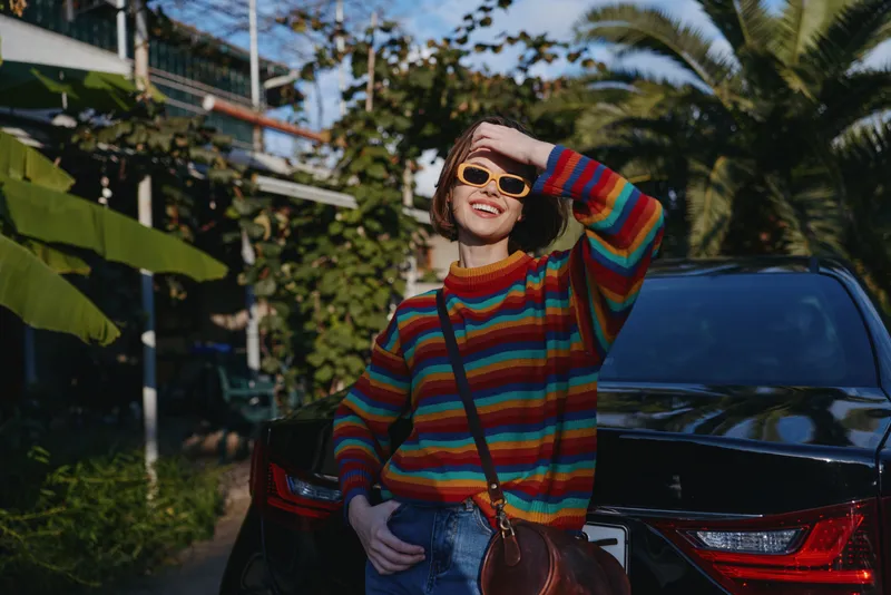 Woman in a striped sweater and sunglasses smiling while leaning on a black car amid tropical plants, short bob haircut, casual outfit and crossbody bag for travel lifestyle portrait.