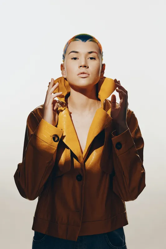 young woman wearing yellow jacket poses with hands near collar, short hair, serious expression, minimal background, fashion portrait bright lighting