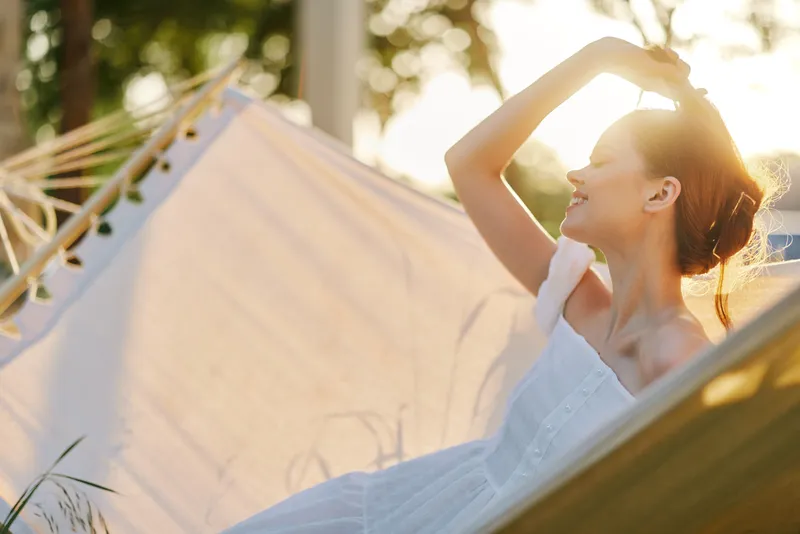 A serene woman in a white dress enjoying a sunny day, feeling relaxed and joyful while lying on a hammock surrounded by nature
