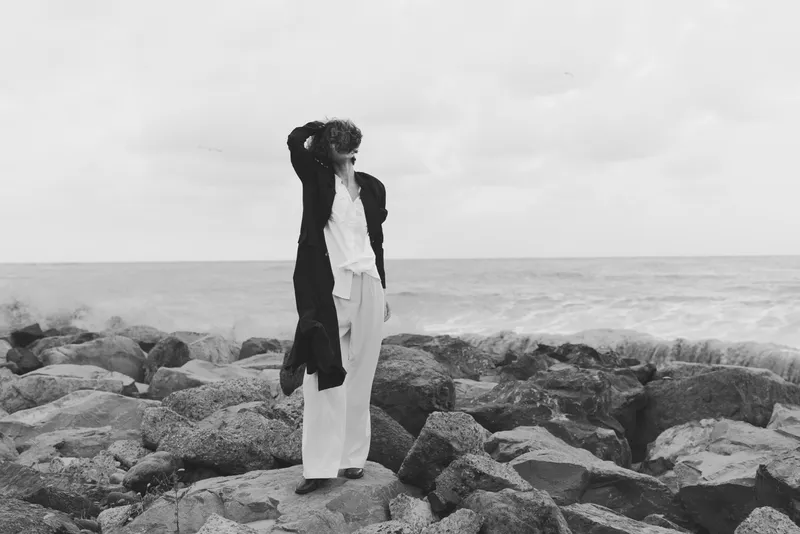 Fashion model stands on rocky shoreline in a white suit and dark coat, posing with hand on head for a moody coastal editorial look and dramatic wind
