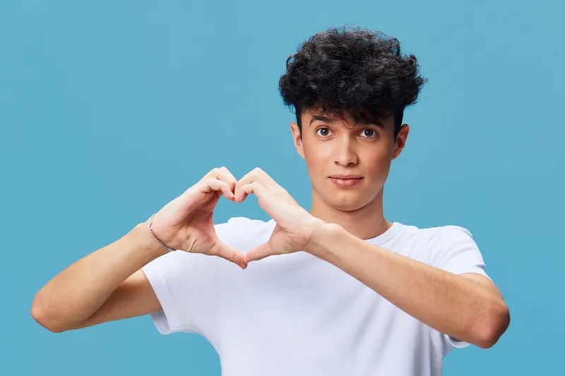 young man making heart shape with hands, curly hair, casual white t-shirt, blue background, friendly expression, isolated