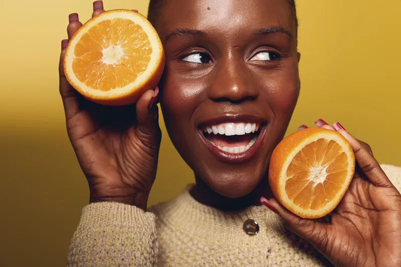 Smiling young black woman with dark skin holding two halves of fresh orange close to her face on yellow background. Concept of healthy lifestyle, vitamin C, and natural beauty.