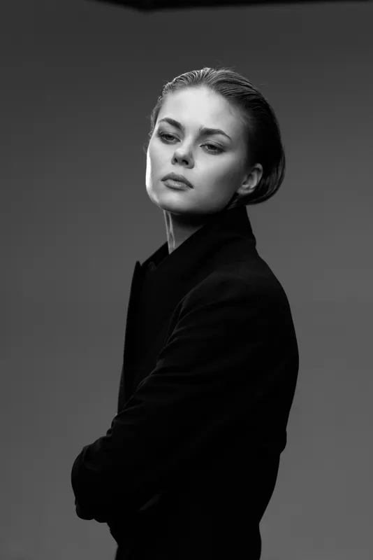 Black and white portrait of a stylish young woman with crossed arms and elegant expression in dark clothing on plain background.