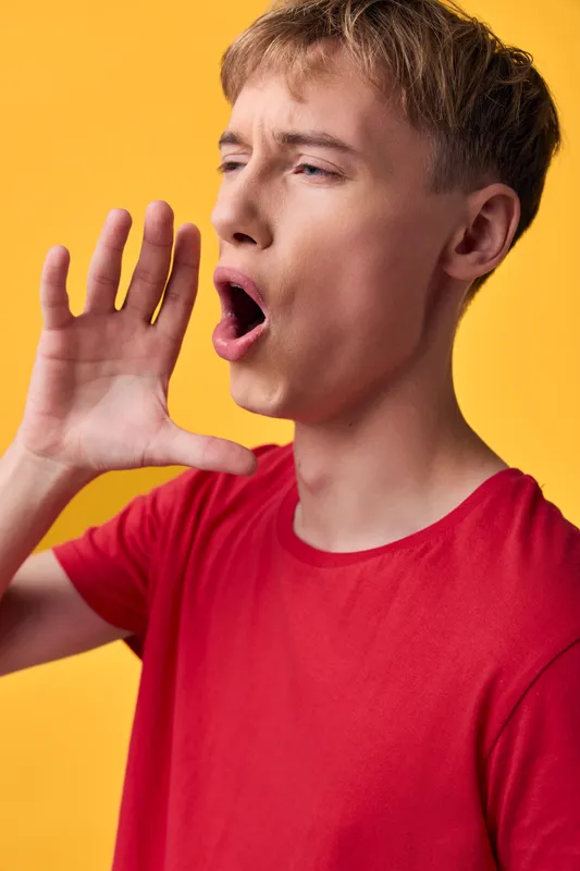 Shouting man with open mouth and hand near his face, wearing a red tee against a bright yellow background, conveying loud expression, energetic communication and attention grabbing gesture for ads