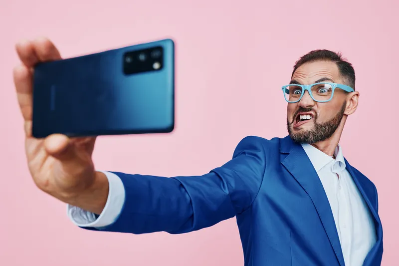 A business professional man takes a dynamic selfie against an isolated pink background, showcasing bold confidence and playful energy in a vivid portrait.