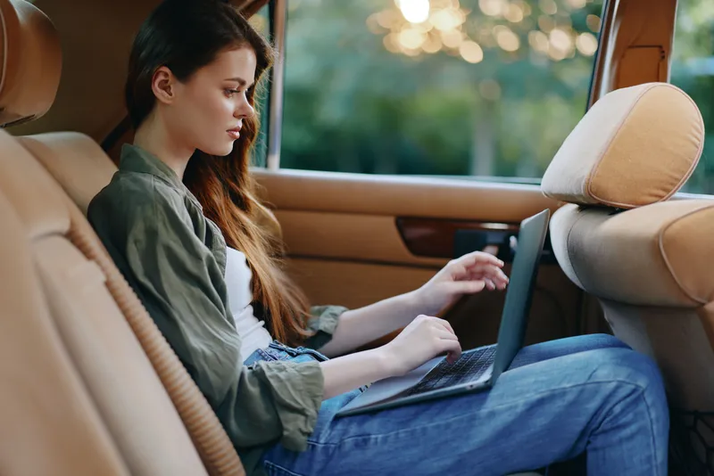 Young woman working on a laptop in a car, wearing casual clothes, focused and engaged, with a green background and warm lighting suggesting a relaxed atmosphere.