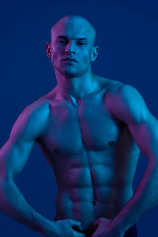 Shirtless man posing under dramatic blue and purple lighting, showcasing strong physique and confident expression in a striking visual composition.