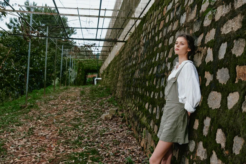 Young woman in a garden wearing a casual olive green dress and white blouse, exuding calmness amidst a lush green background, ideal for nature and fashion themes