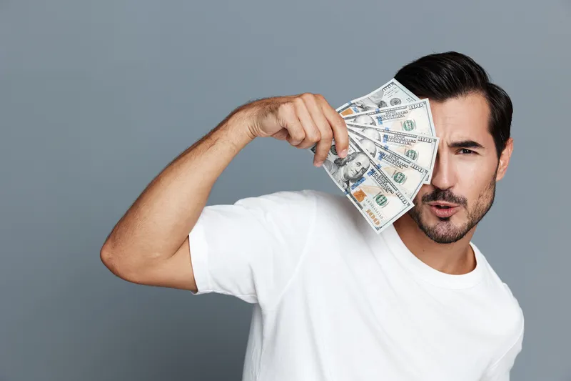 Handsome man holding fan of cash covering one eye, looking confident and playful towards the camera. Studio portrait with neutral grey background, financial success concept.