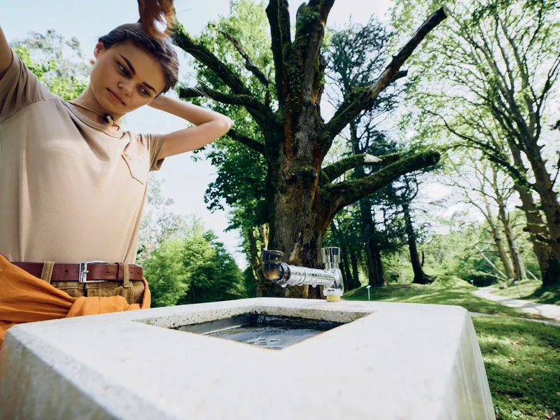 Woman by outdoor fountain in park tying hair above a stone basin, casual portrait with large tree and lush greenery, summer nature scene with water and relaxed atmosphere.