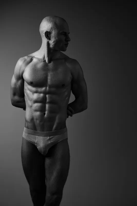 Black and white image of a muscular man showcasing his physique, emphasizing strength and fitness in a dramatic pose against a soft gradient background.
