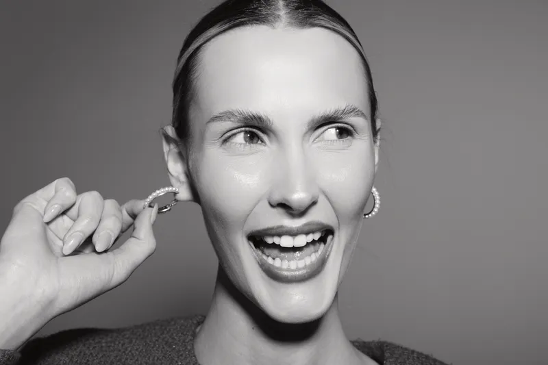 Smiling white woman with light skin and slicked hair adjusts hoop earring, expressing joy and confidence in close-up portrait.