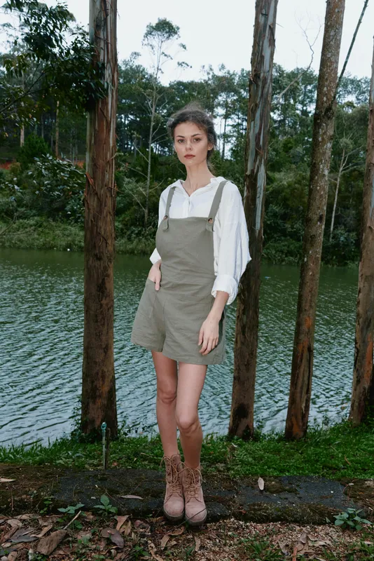 Stylish young model in olive green overalls and white blouse posing by a tranquil lake surrounded by trees, showcasing a natural setting and casual outdoor fashion