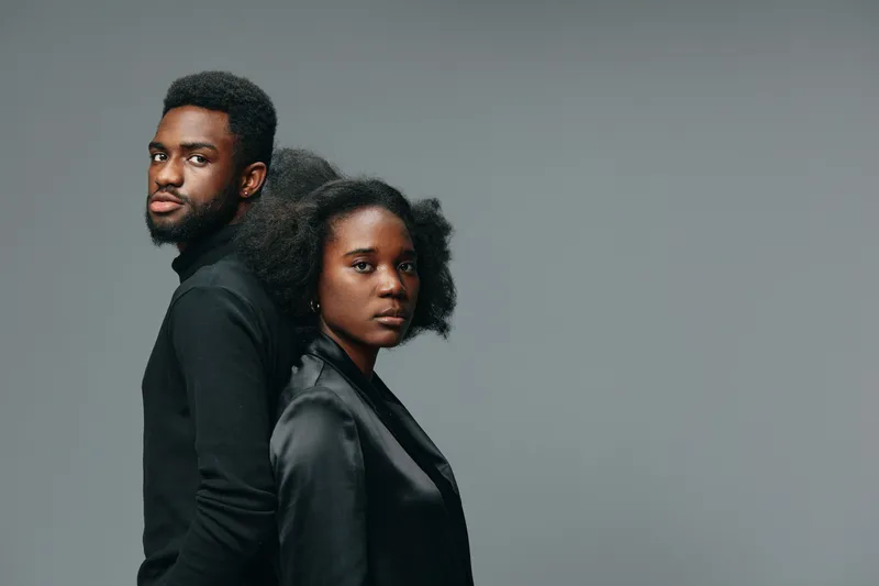 Portrait of a stylish black couple standing back to back, showcasing confidence in modern attire against a minimalist gray background