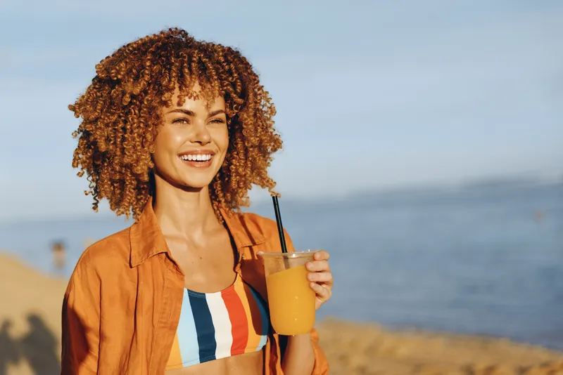 A woman with curly hair shows smile while holding iced coffee cup on a sunny beach, ocean horizon softly blurred behind, outdoor summer vacation mood, warm sunlight and sandy coast