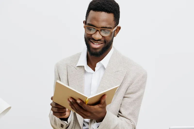 Cheerful smiling man reading book with glasses, wearing beige blazer and white shirt, standing against plain light background. Casual clothing, positive mood, learning concept, modern style, indoor st