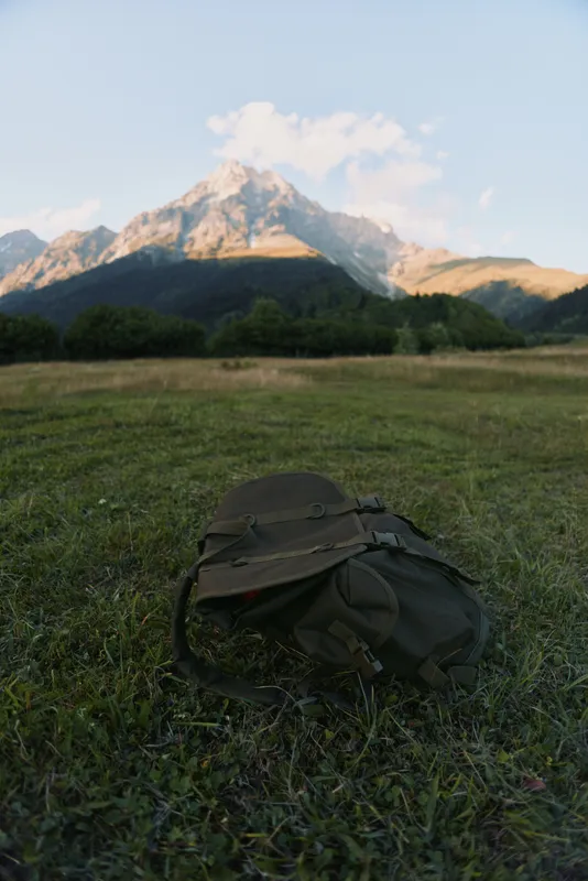 Backpack resting on a grassy field with a dramatic mountain range in the background, conveying outdoor travel, nature, and exploration vibes, conveying freedom, adventure.