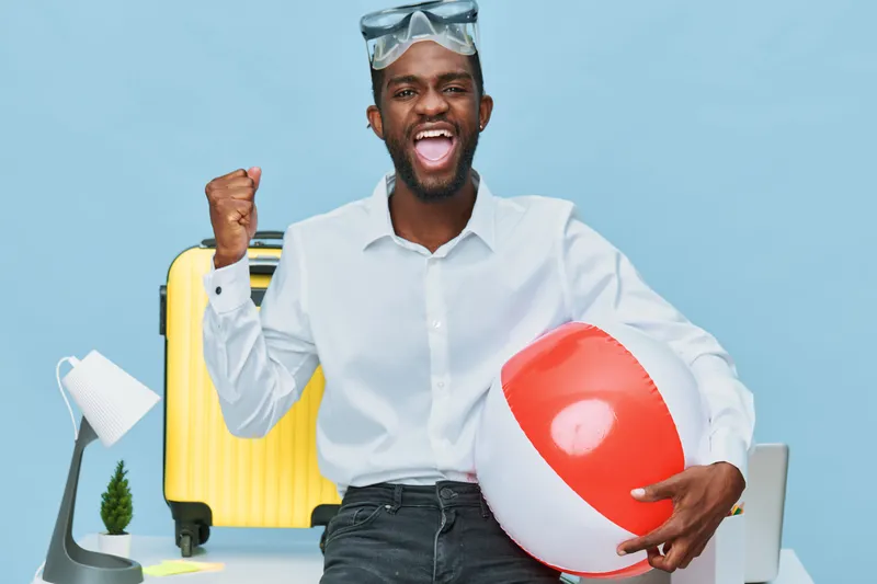 Smiling man with dark skin, wearing white shirt and sunglasses on head, holding a red and white beach ball, surrounded by travel accessories like a yellow suitcase and lamp, in casual vacation setting