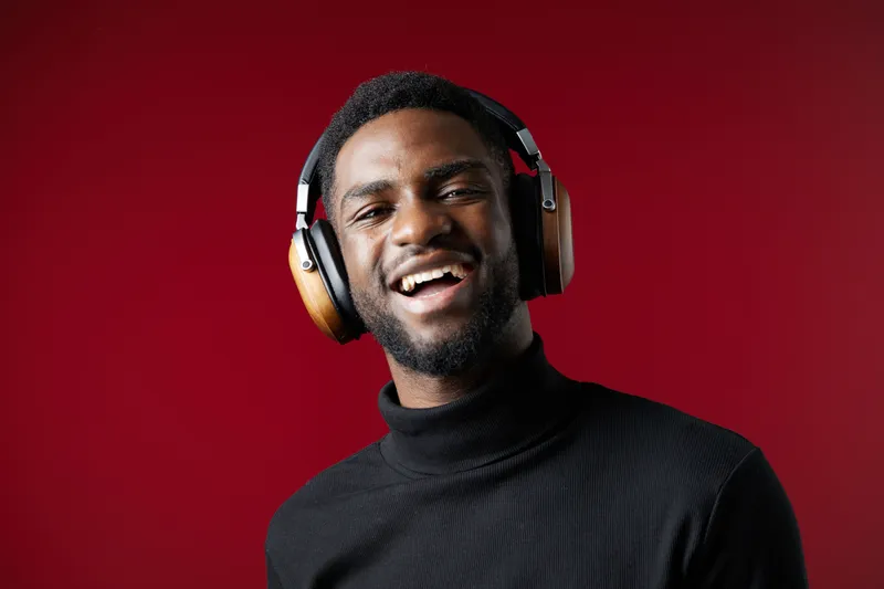 Happy young Black man in a stylish black turtleneck and headphones, smiling joyfully against a vibrant red background, embodying music enjoyment and modern lifestyle