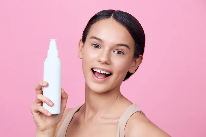 Young woman holding a white spray bottle against a pink background, smiling, showcasing skincare, beauty, and healthy lifestyle concepts, perfect for beauty marketing