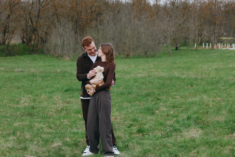 A couple outdoors with a goat kid in a grassy park, showcasing affection and a lifestyle moment with animals. A warm portrait of love, nature, and calm togetherness in a serene field setting.