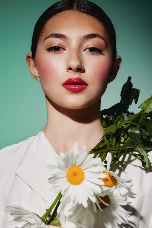Young woman with vibrant red lipstick and bold blush holds fresh daisies against a teal background. Fashion and beauty concept with creative floral accessories and contemporary elegance.