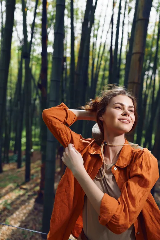 Woman smile bamboo forest portrait outdoor, young female in orange jacket enjoying sunlight in bamboo grove, relaxed expression, natural beauty and travel lifestyle scene.