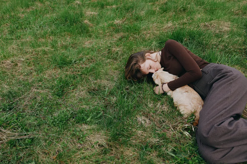 A woman lies on grass embracing a young goat kid in a peaceful outdoor scene. Warm light, relaxed mood, countryside ambiance, gentle companionship, natural textures convey a lifestyle moment of