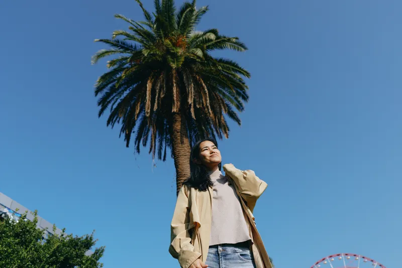 Woman under a tall palm in a sunny outdoor scene wearing a beige jacket and denim shorts, relaxed pose against a clear blue sky and vibrant daylight and warmth.