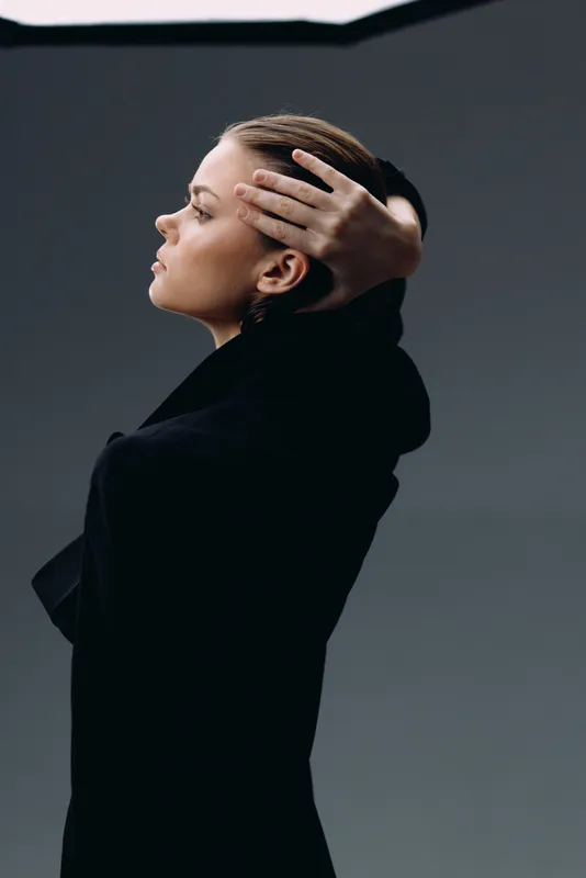 Side profile of a thoughtful woman with a hand on her head wearing black clothes in a dark background.