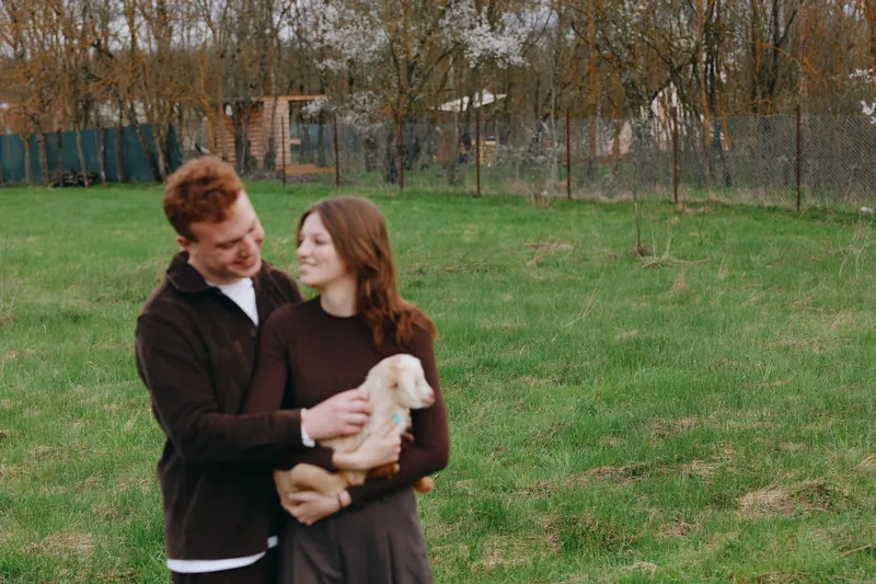 A young couple in a field embrace a light puppy, smiling softly as they lean close. The scene conveys simple joy, companionship, and rural lifestyle, capturing intimate connection and animal
