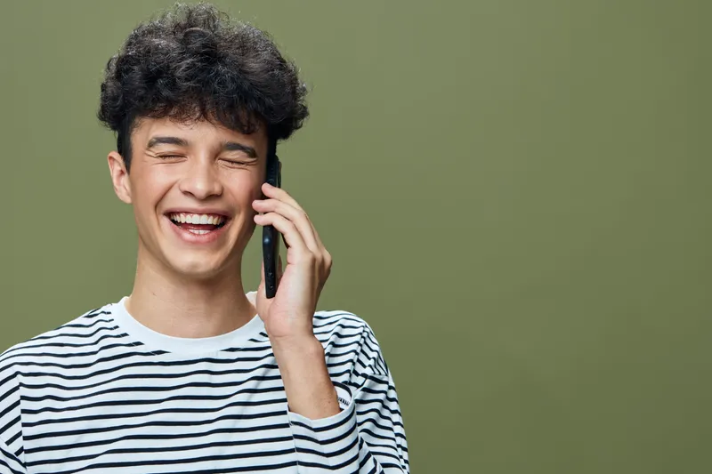smiling man talking on smartphone with striped shirt against green background, cheerful young adult enjoying phone call in casual style