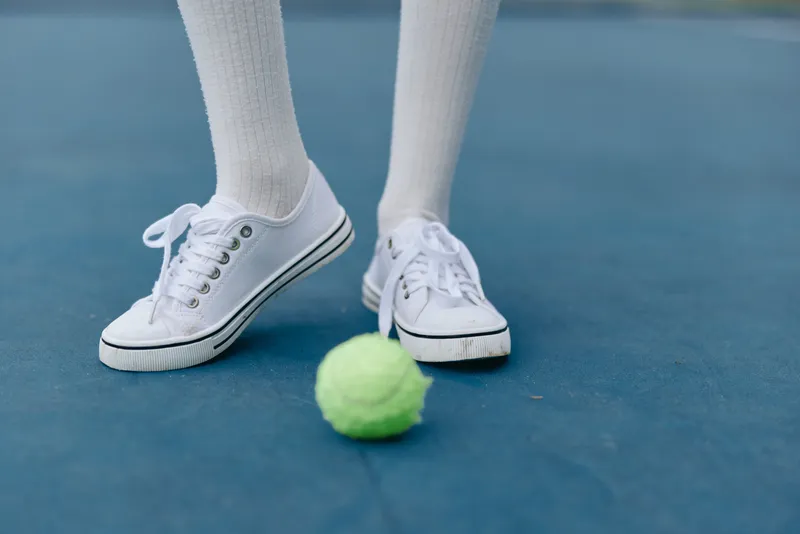 Young athlete in white sneakers and socks standing on a tennis court with a bright green tennis ball, capturing the excitement of sports and teamwork