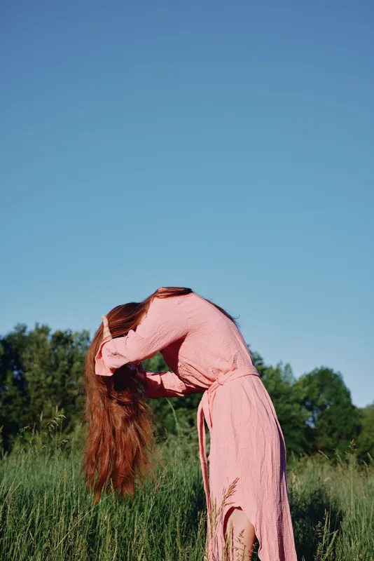 Woman in a pink dress embracing nature by brushing her long hair in a lush green field under a clear blue sky, conveying serenity and freedom