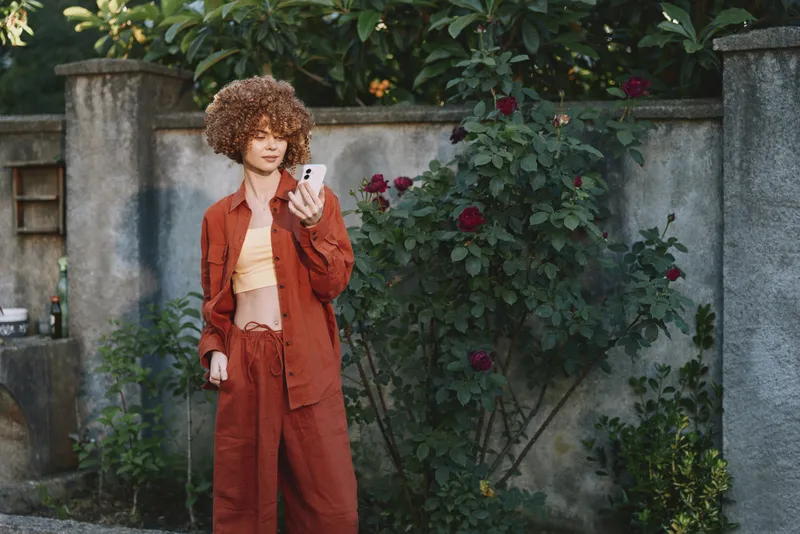 Vibrant woman outdoors wearing rust suit and crop top, checking her phone, relaxed pose, sunlit garden backdrop, natural colors, candid lifestyle portrait moment