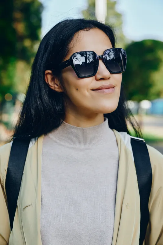 Sunglasses worn by a casual woman outdoors on a sunny day, light jacket and backpack, urban street style with a relaxed smile, modern chic vibe in a park setting
