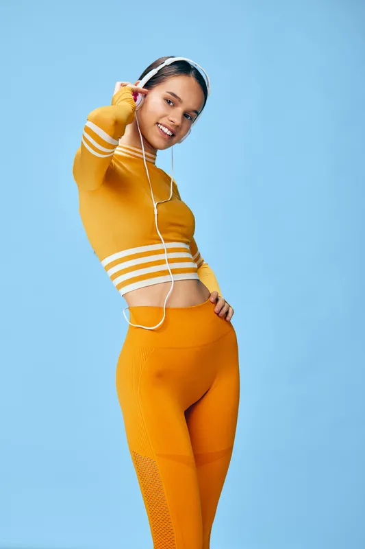 Young woman fitness portrait in bright orange crop top and leggings, headphones on, smiling and stretching in a sport pose against a clean light blue studio background.