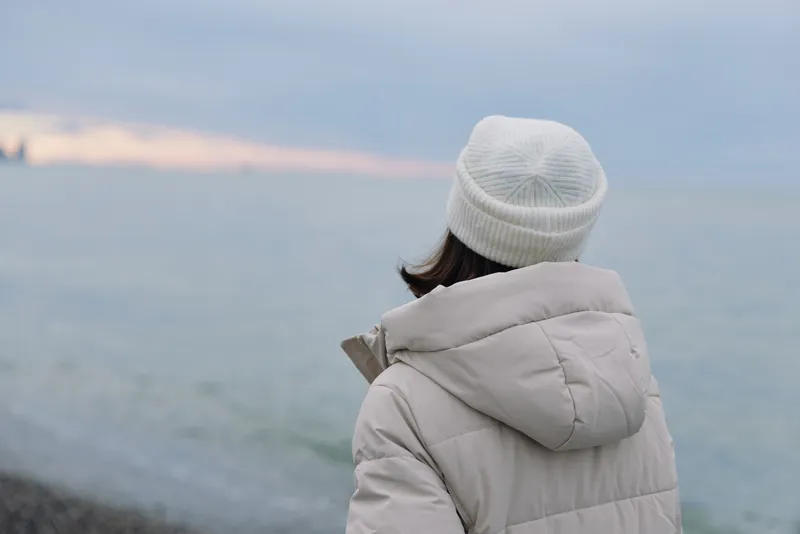 Person in a warm winter coat and beanie gazes at a calm sea along a quiet coast, capturing a tranquil moment of reflection and distant horizons under soft lighting.