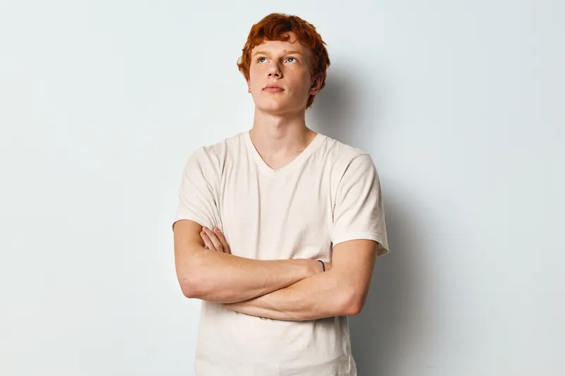 Young man with red hair wearing beige t shirt standing with arms crossed against light background looking away thoughtfully with confident expression and casual posture.